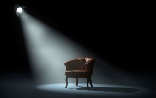 A single armchair lit by a theatrical spotlight in a darkened space